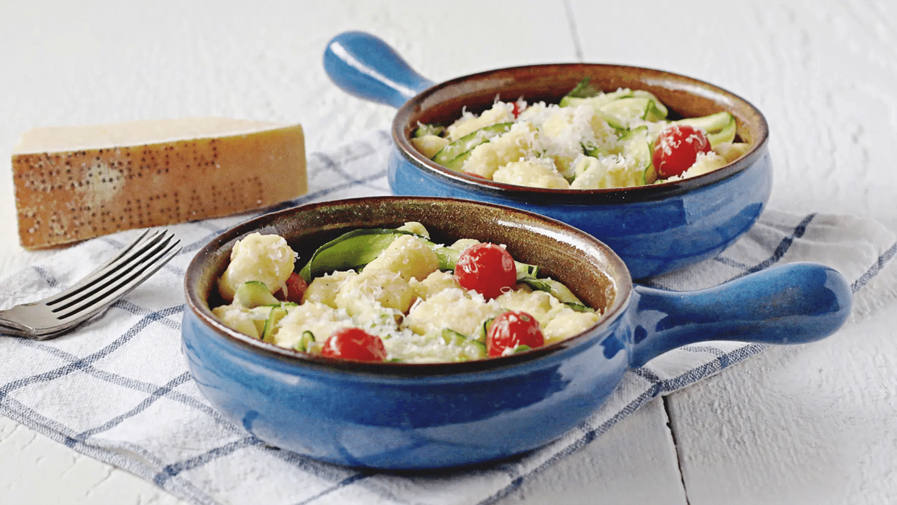 Two bowls with gnocchi, zucchini, and tomatoes on tablecloth.