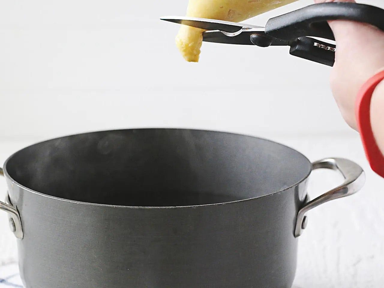 Scissors cutting Ziploc bag of gnocchi dough over cooking pot.