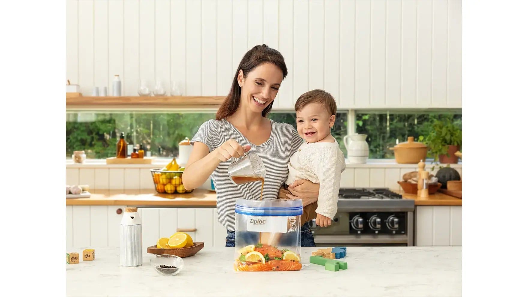Woman holding a baby and pouring marinade into a Ziploc bag of salmon.