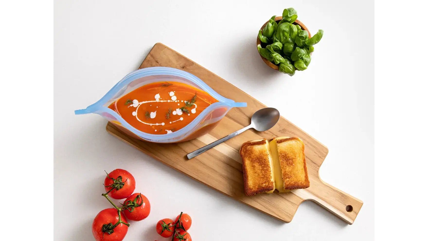 Tomato soup in Ziploc Endurables container with grilled cheese on cutting board.